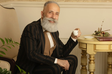 Senior handsome concentrated man with beard in business suit sitting and drinking coffee.
