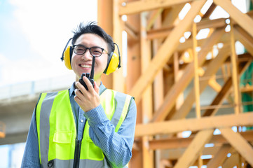Young asian engineers are working on the construction site. Wear a yellow earmuff sound protection and uniform staff. Hand holding portable radio transceiver for communication - obrazy, fototapety, plakaty