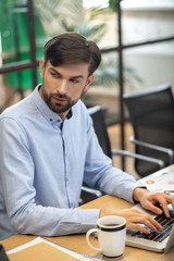 Young bearded man in a blue shirt looking serious while working on a laptop