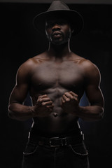 Fototapeta na wymiar portrait of a handsome black man in dark hat and jeans with naked sports torso looking in the camera on dark background and holds his fists in front of him