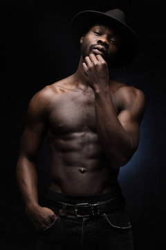 portrait of a handsome black man in dark hat and jeans with naked sports torso looking in the camera on dark background
