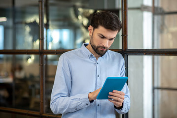 Young bearded man in a blue shirt standing and looking at the tablet
