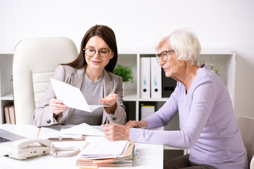 Fototapeta premium Smiling young social worker sitting at table with senior lady and explaining document content to her