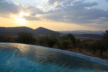 zachód słońca nad basenem typu infinity pool z naturalnym afrykańskim krajobrazem w tle - obrazy, fototapety, plakaty