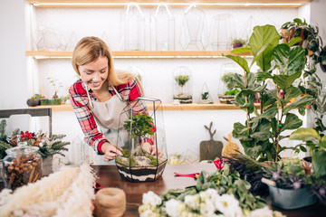 young beautiful lady florist at her own floral shop taking care of flowers, wearing red casual shirt and apron, female with blond hair surrounded by green plants