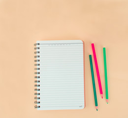 Three color pencils scattered beside a white notebook on a light orange background paper.