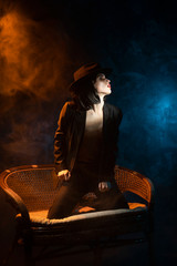 Beautiful braless girl, wearing a unbuttoned black blazer, brunette wig and hat, sensually posing on sofa on dark background next to the blue and warm light lamp in the theatrical smoke. Noir design.