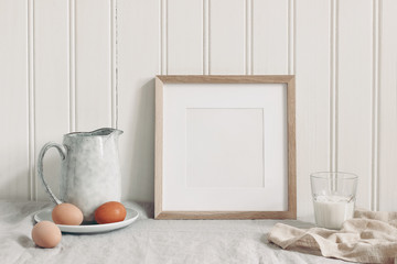 Spring breakfast still life scene. Square empty wooden frame mockup with chicken eggs,, glass of milk and ceramic jug. Easter food and drink concept. Farmhouse, Scandinavian design.