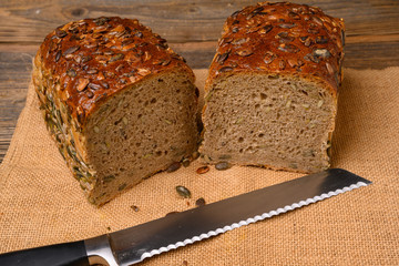 A fresh pumpkin seed bread from the baker halved with a bread knife on a jute fabric on a rustic wooden background.