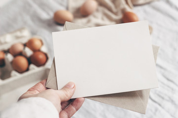 Closeup of woman's hand holding blank paper card. Greeting card mock-up scene. Spring, Easter design. Feminine styled stock photo. Blurred background with chicken eggs and line tablecloth. Top view.