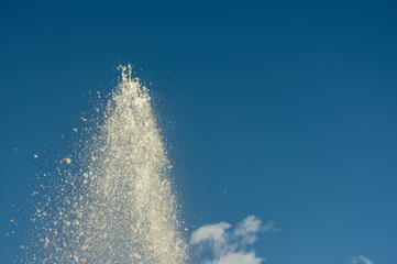 Spitze einer Wasserfontäne eines Springbrunnens bei strahlendem Sonnenschein gegen blauen Himmel mit kleinen weißen Wolken