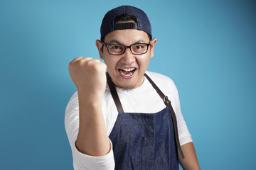 Portrait of happy excited proud Asian chef or waiter shows winning gesture, against blue background