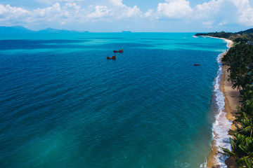 Obraz premium Aerial scenery view of white sand beach shore with turquoise clear sea water and foamy waves. Bird's eye panoramic view of picturesque coastline bay with big fishermen boats. Samui island, Thailand