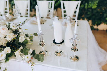 A wedding table with a white tablecloth, candles, a large bouquet of white roses on a background of green foliage of wild grapes. Outdoor, summer ceremony. Close up.