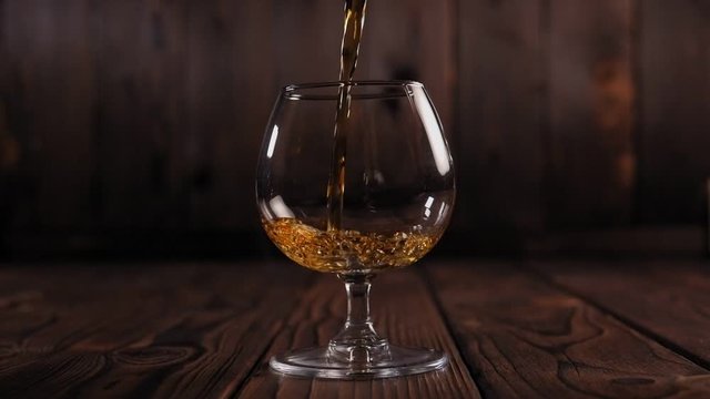 Pouring brandy or cognac from the bottle into the glass against wooden background. Cognac is poured from a bottle into a glass on a wooden background. Slow motion.