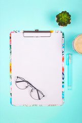 Blank paper on colored clipboard with copy space on a bright neo-mint background with glasses and succulents. Top view, flat lay.