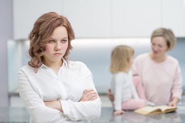 A teenage girl looks upset at the camera, in the background her mother is playing with her younger sister. Problems of puberty, depression, problems in the family