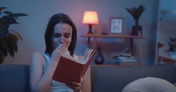 Front view of young upset girl with tears on her face reading book. Millennial brunnette woman sitting on sofa crying because of book plot and using paper tissue.Home background.