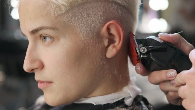 The hairdresser cuts blonde female client with a trimmer. A short pixie haircut and shaved temples . Ornament on the hair.