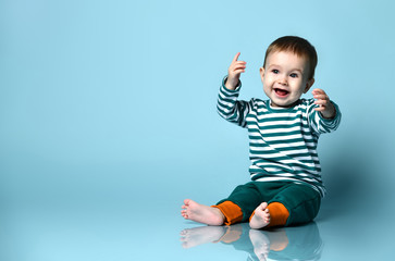 Little baby boy in stylish casual clothing barefoot sitting on floor and smiling with raised hands over blue wall background - obrazy, fototapety, plakaty