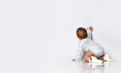 Baby girl in gray bodysuit and socks with red bows. She is creeping on the floor, touching wall, isolated on white. Back view
