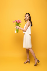 full length view of attractive young woman smiling at camera while holding bouquet of pink tulips on yellow background
