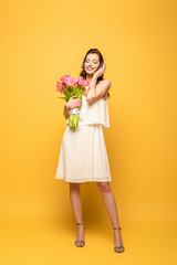 full length view of happy young woman in wireless headphones holding bouquet of pink tulips on yellow background