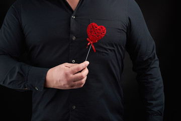 adult man stands on a dark background wearing a black shirt and holding a red carved heart