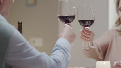 Tracking selective focus shot of cheerful elderly woman clinking glasses in toast with husband and drinking red wine at romantic dinner - Powered by Adobe