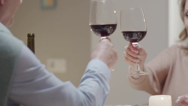 Tracking selective focus shot of cheerful elderly woman clinking glasses in toast with husband and drinking red wine at romantic dinner