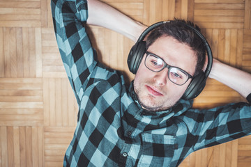 Enjoying music at home: Young Caucasian man is lying on the wooden floor and listening to music...