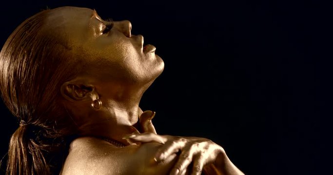 close-up of a female face with Golden skin in profile on a black background. the woman closed her eyes, lifted her head up, and slowly slid her fingers along her neck