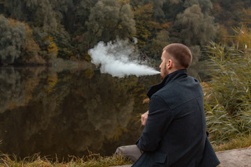 A guy with a beard sits on the lake and smokes an electronic cigarette. Blows puffs of smoke. The concept of unity with nature. Rest near the water. Vape, Man Vaping. lips and smoke, a lot of smoke.