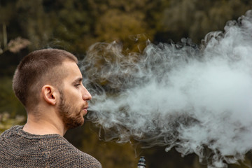 A guy with a beard sits on the lake and smokes an electronic cigarette. Blows puffs of smoke. The concept of unity with nature. Rest near the water. Vape, Man Vaping. lips and smoke, a lot of smoke.