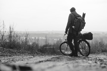 Fototapeta na wymiar A rider man with a backpack and a bicycle stands and looks into the distance. hilltop overlooking a valley in haze, a city on the horizon. winter or autumn landscape road. Black and white