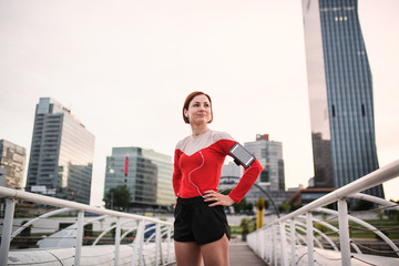 Young woman with smartphone standing outdoors in city, resting after exercise.