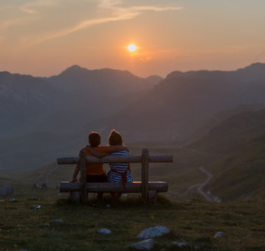Boy and a girl sit hugging each other on a bench and watch the sunset nere winding road seen from Sedlo Pass in Durmitor National Park in Montenegro at sunset