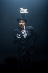 emotional magician in hat with dove holding wand in dark room with smoke - obrazy, fototapety, plakaty