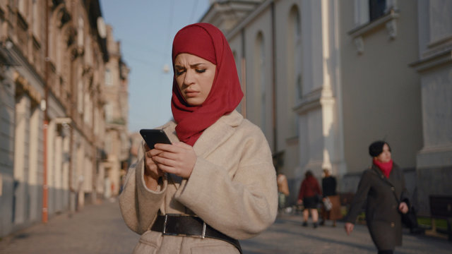 Sociable busy young arab woman messaging on a smartphone outdoors. Portrait of worried pretty Muslim girl in hijab looking at cellphone.