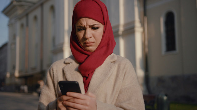 Portrait of embarassed young Muslim woman looking at smartphone. Surprised arab girl on the busy street. Shocking news.