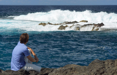 Fototapeta na wymiar Young man sitting on rock shore and looking at ocean. View from behind. Blue ocean with high waves, water splash