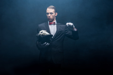 emotional magician in suit showing trick with wand and white bunny in hat, dark room with smoke