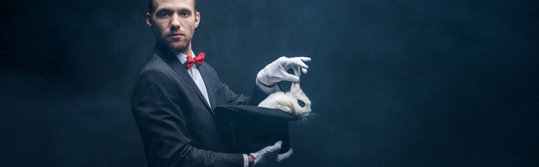 panoramic shot of young magician in suit showing trick with white rabbit in hat, dark room with...