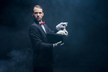 professional magician in suit showing trick with white rabbit in hat, dark room with smoke