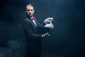 Fotobehang professional magician in suit showing trick with white rabbit in hat, dark room with smoke © LIGHTFIELD STUDIOS