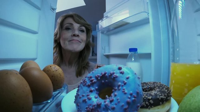 View from inside of refrigerator of senior woman opening door, taking blue glazed donut from shelve and eating it with pleasure in the night at home