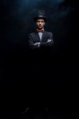 magician in suit and hat standing with crossed arms in dark smoky room - obrazy, fototapety, plakaty