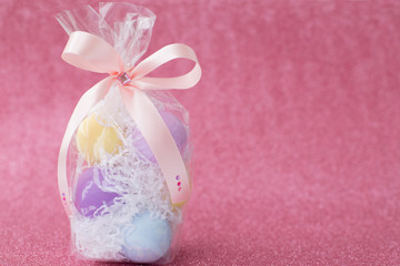 Multi-colored purple, pink, blue, yellow Easter eggs in a transparent gift package, decorated with a bow and rhinestones, on a pink shiny bokeh background. Symbol of celebration of religious holiday