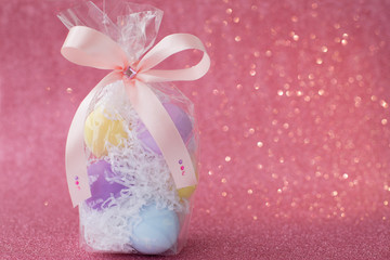 Multi-colored purple, pink, blue, yellow Easter eggs in a transparent gift package, decorated with a bow and rhinestones, on a pink shiny bokeh background. Symbol of celebration of religious holiday