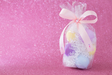 Multi-colored purple, pink, blue, yellow Easter eggs in a transparent gift package, decorated with a bow and rhinestones, on a pink shiny bokeh background. Symbol of celebration of religious holiday